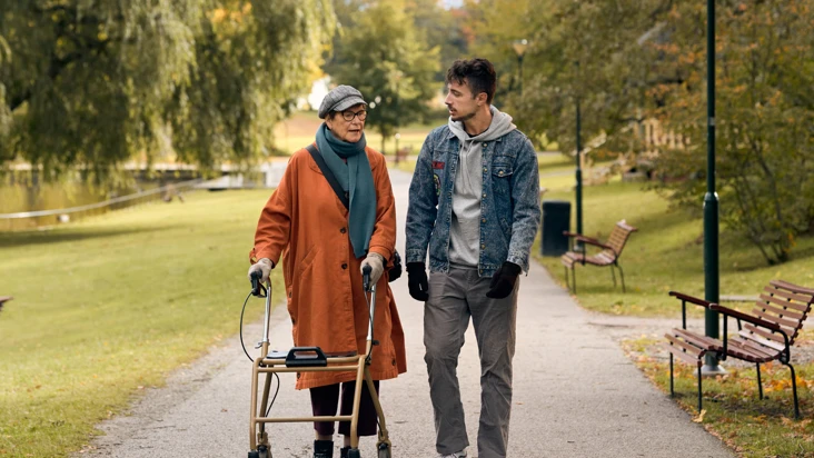 En äldre kvinna med rullator promenerar i en park tillsammans med en ung man.