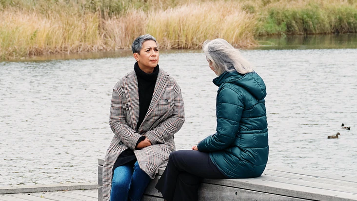Två kvinnor sitter på en brygga vid en sjö och samtalar allvarligt.
