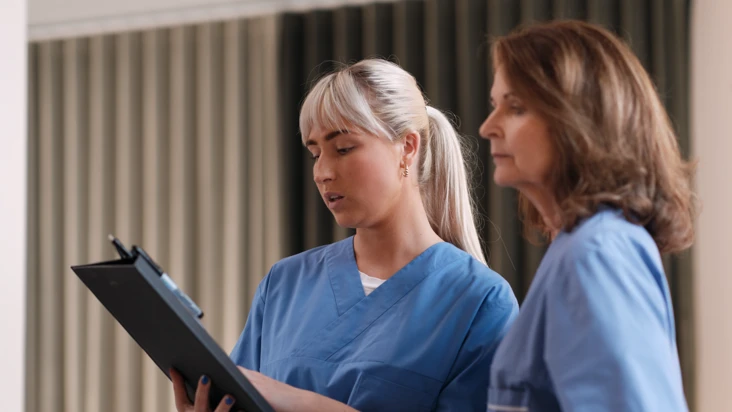 Två kvinnliga kollegor i blå vårdkläder tittar tillsammans på ett papper.