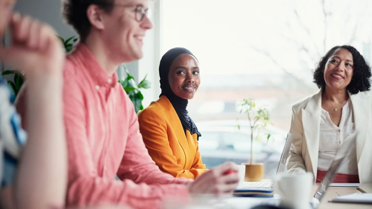 Tre personer har m&ouml;te runt ett kontorsbord i ett kontorsrum. De tittar p&aring; varandra, samtalar och ler
